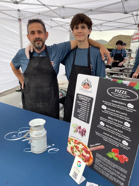 Ferruccio's founder and his son inside the mobile pizza catering tent beside the menu board