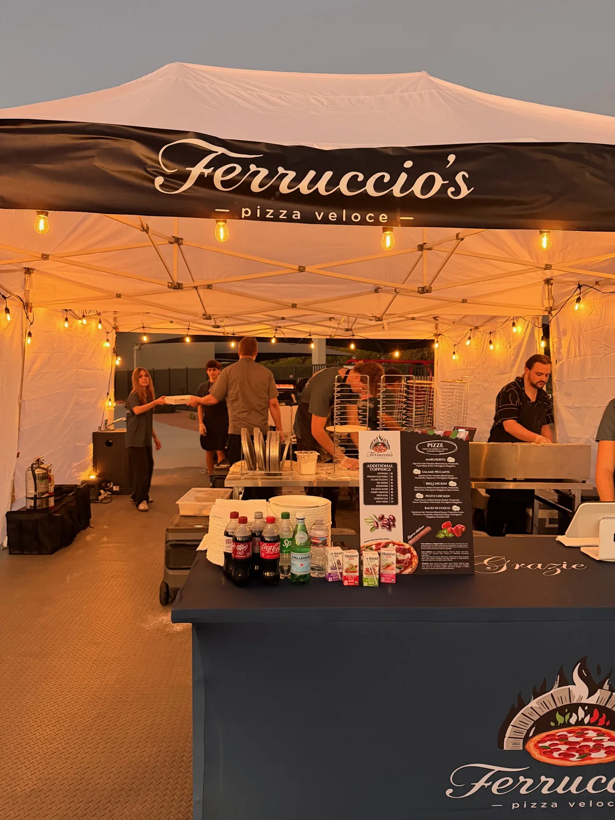 Ferruccio's tent glowing with string lights at an evening event while the team prepares pizzas